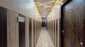 Un pasillo con piso de diseño geométrico, una puerta de madera con placa de número y un techo con iluminación lineal.