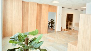A spacious room with wooden panel walls, a large plant in the foreground, and a view into another room with a potted plant and a sofa. - Alpenruhe Vacation Rental with Exceptional Mountain Views, Pool, and Sauna (Scheidegg)