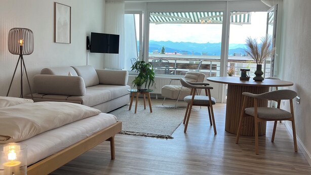 A modern living room with a sofa, a coffee table, and a dining area with chairs. There is a TV mounted on the wall and a view of mountains through large windows. - Alpenruhe Vacation Rental with Exceptional Mountain Views, Pool, and Sauna (Scheidegg)