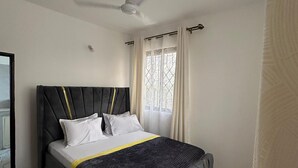 Une chambre à coucher avec un lit, un ventilateur de plafond, une fenêtre avec des rideaux et un sol carrelé.