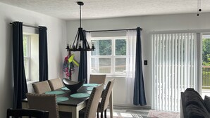 Ein Essbereich mit einem Holztisch, Stühlen und einer Pendelleuchte. Ein Fenster mit Vorhängen bietet Blick auf Grünflächen.