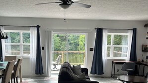 Ein geräumiges Wohnzimmer mit einem dunklen Sofa, einem runden Tisch und einem Deckenventilator. Große Fenster mit Vorhängen sorgen für natürliches Licht und einen Blick ins Freie.
