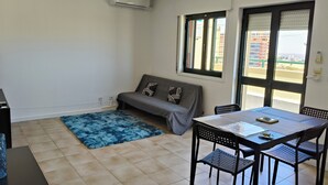A room with a sofa, a table with chairs, a blue rug, and an air conditioning unit.