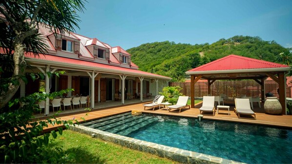 A pool area with lounge chairs and a gazebo, surrounded by a house with a red roof and a covered patio.