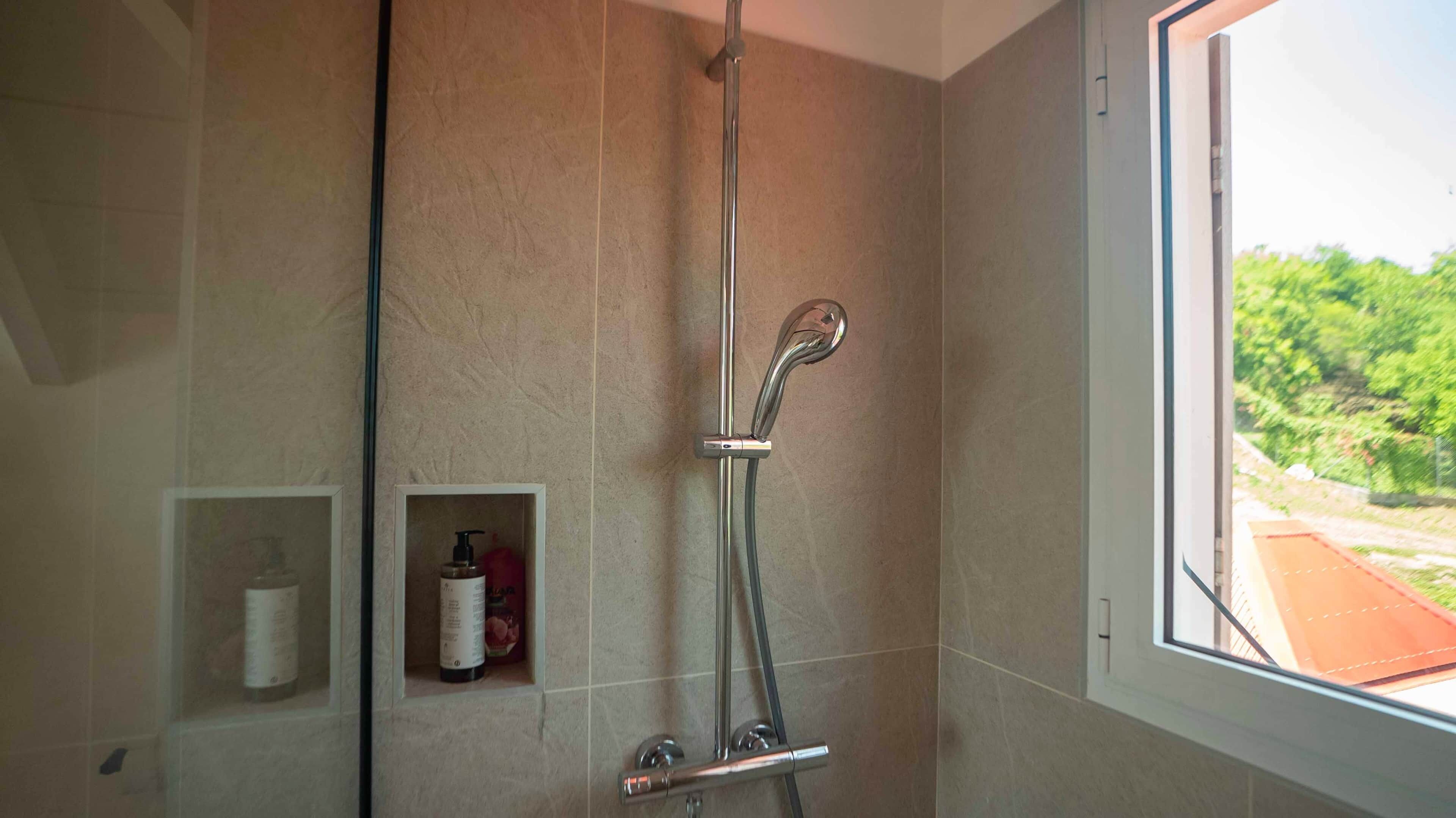 A shower with a glass door, a small shelf with toiletries, and a window with a view of greenery.
