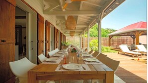 A covered dining area with a long wooden table set for a meal, surrounded by white chairs.