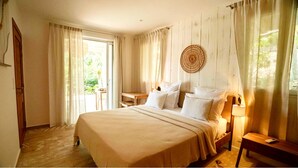 A bedroom with a large bed, a bedside table, a lamp, and a view of greenery through the window.