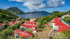 A coastal village with red-roofed houses, a marina with boats, and a mountainous backdrop.