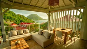 A balcony with a sofa, table, and chairs, overlooking a scenic view of a mountain and water.