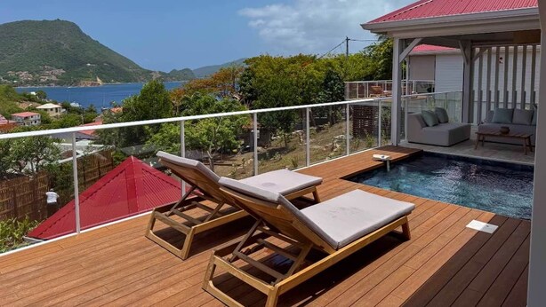 Une terrasse en bois avec des chaises longues et une piscine, offrant une vue sur un paysage montagneux et la mer.