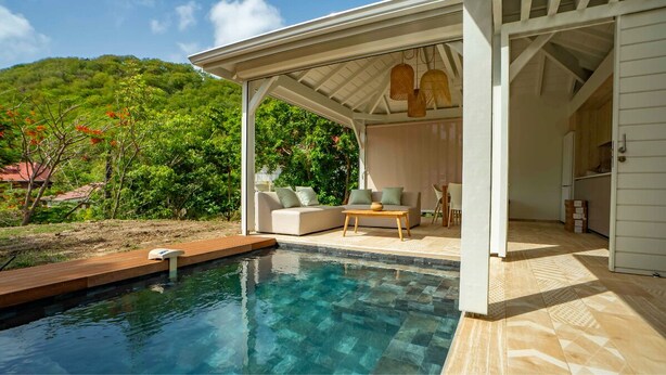 Un espace aménagé au bord de la piscine, comprenant un coin salon couvert, une terrasse en bois et une vue sur une colline verdoyante.