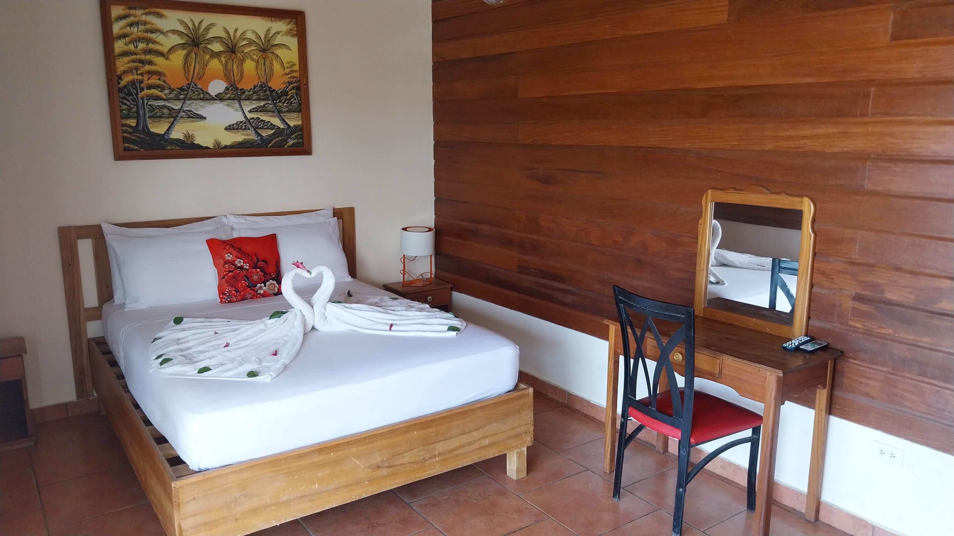 Una cama bien hecha con un almohadón rojo, un escritorio de madera con un espejo, y un cuadro con un atardecer sobre palmeras en la pared.