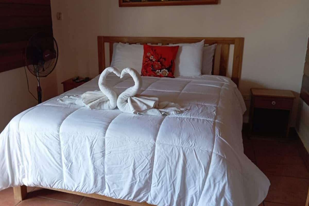 A bed with a white quilt and two swan-shaped pillows, a red pillow, a wooden headboard, a bedside table, and a fan.