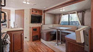 Een compact interieur van een camper met een kitchenette, een flatscreen-tv, een eethoek en een raam met gordijnen.