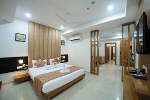 A modern hotel room with a large bed, bedside tables, a flat-screen TV, an air conditioning unit, and a hallway leading to another room.