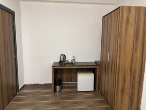 Una habitación pequeña con un escritorio de madera, una tetera, una botella de agua, un cubo de basura y un refrigerador pequeño.