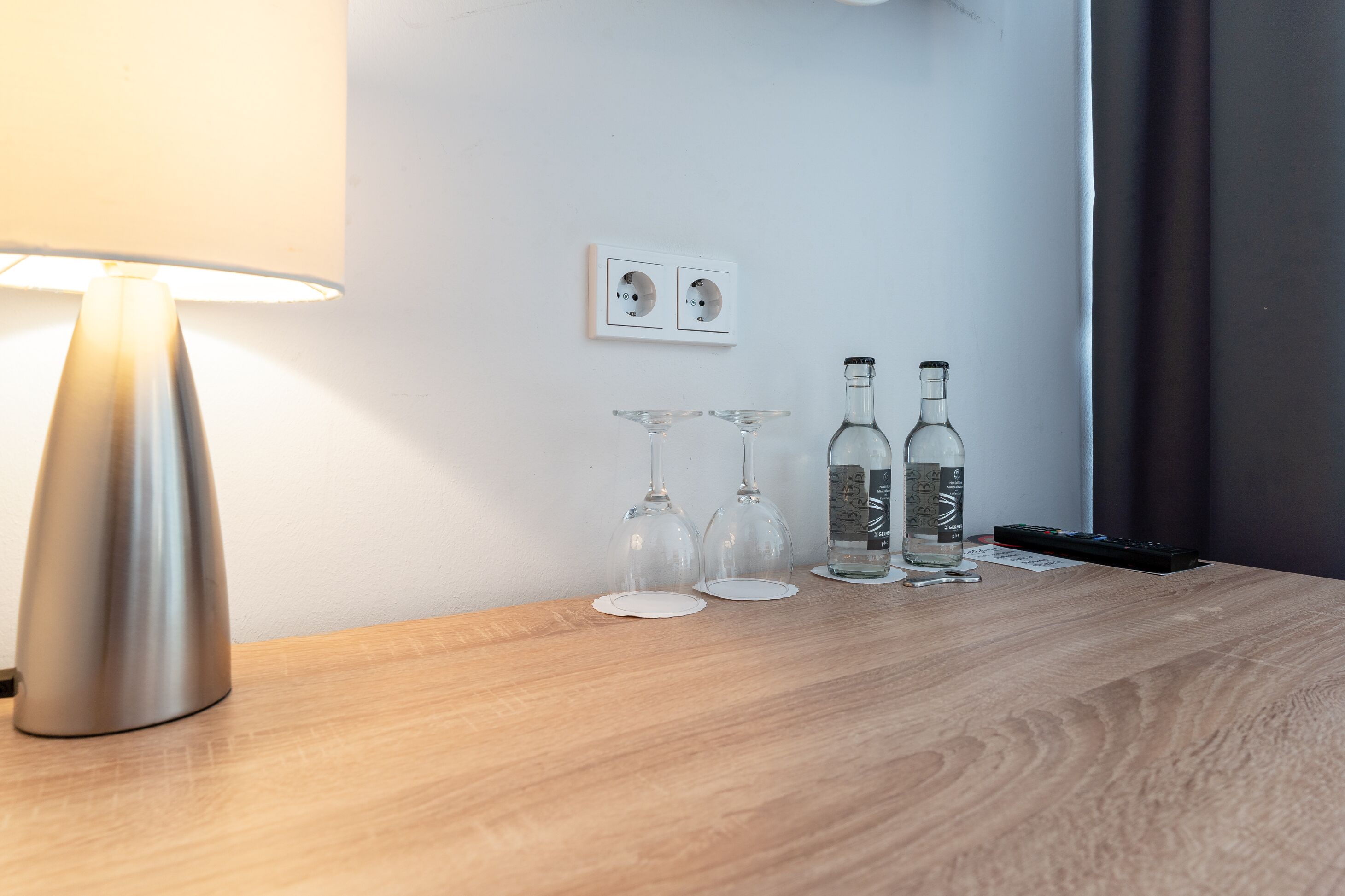 A wooden table with a lamp, two wine glasses, and two bottles.