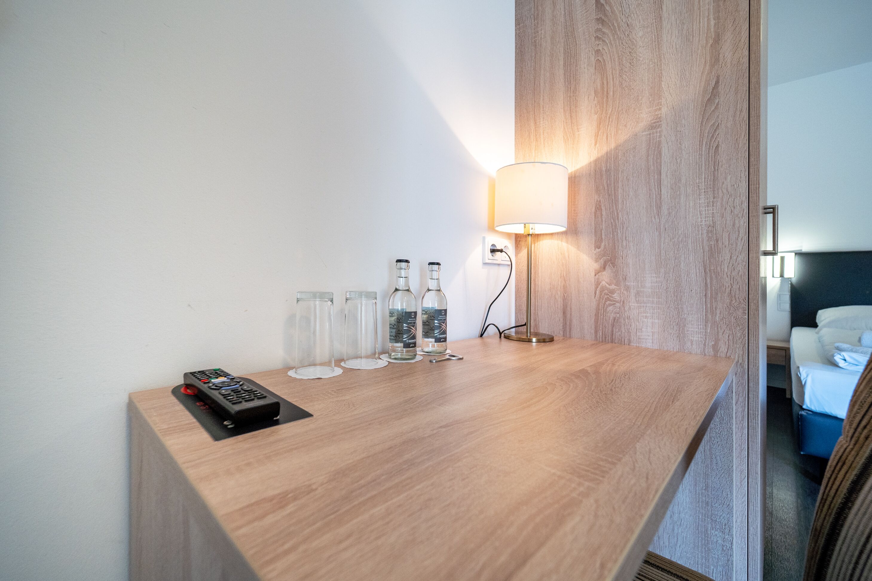 Un escritorio de madera con un control remoto, tres botellas de vidrio y una lámpara de pared en una habitación de hotel.