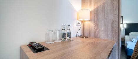 Un escritorio de madera con un control remoto, tres botellas de vidrio y una lámpara de pared en una habitación de hotel.