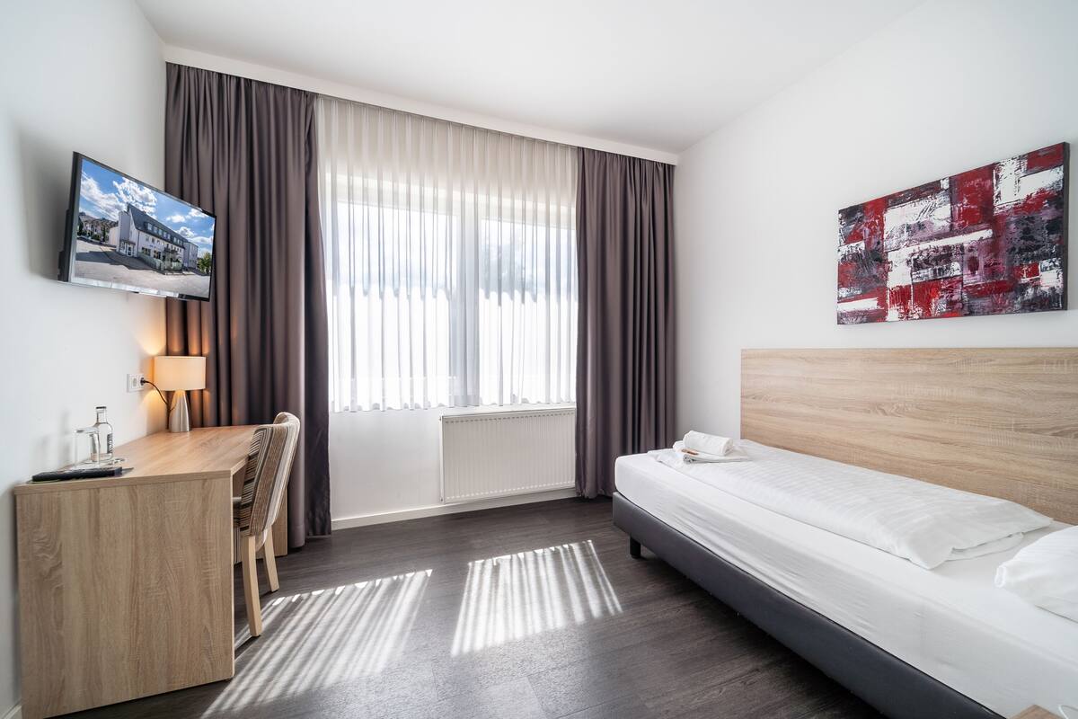 Hotelkamer met een bed, een bureau met stoel, een aan de muur bevestigde televisie en een raam met gordijnen.