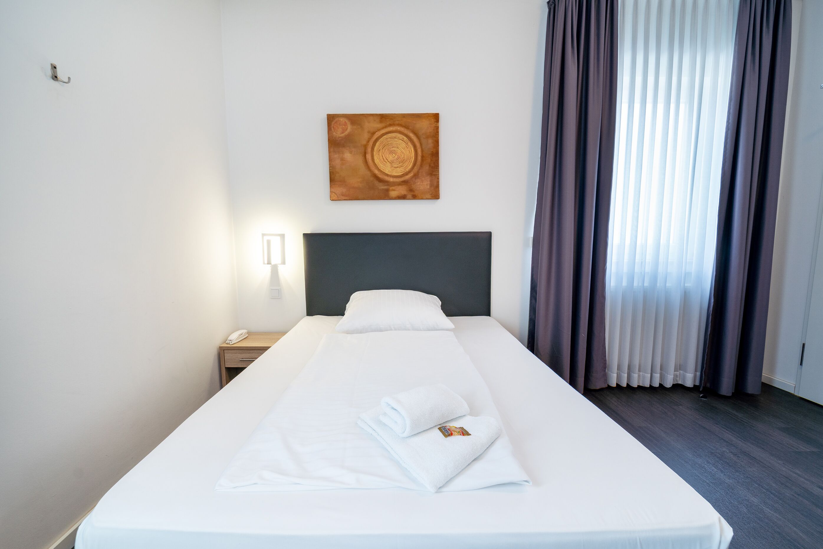 Una cama bien hecha con sábanas blancas y una toalla doblada, una mesita de noche con un teléfono, una obra de arte en la pared y una ventana con cortinas.