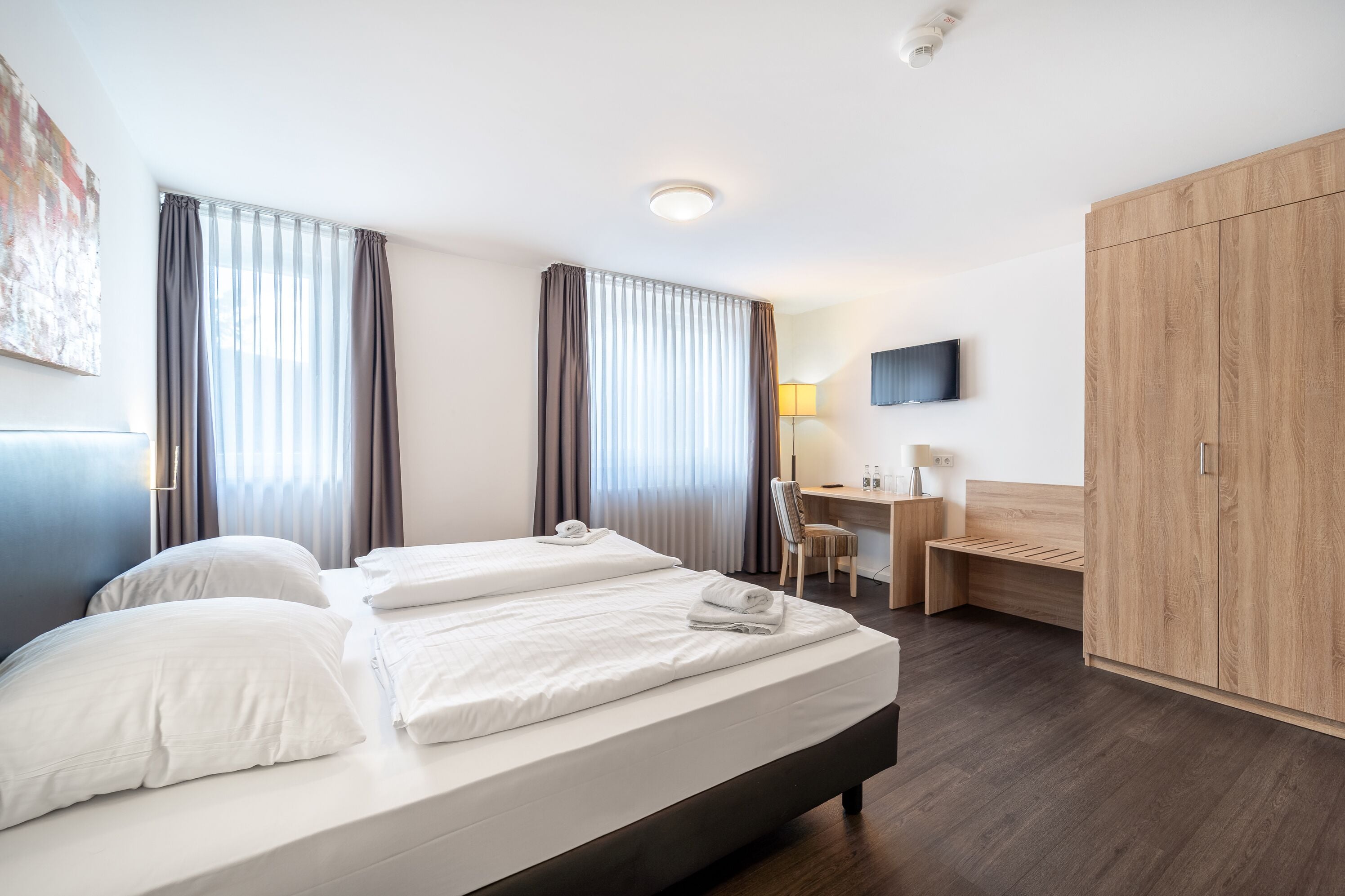 A hotel room with a large bed, a desk, a chair, a wardrobe, and a TV on the wall.