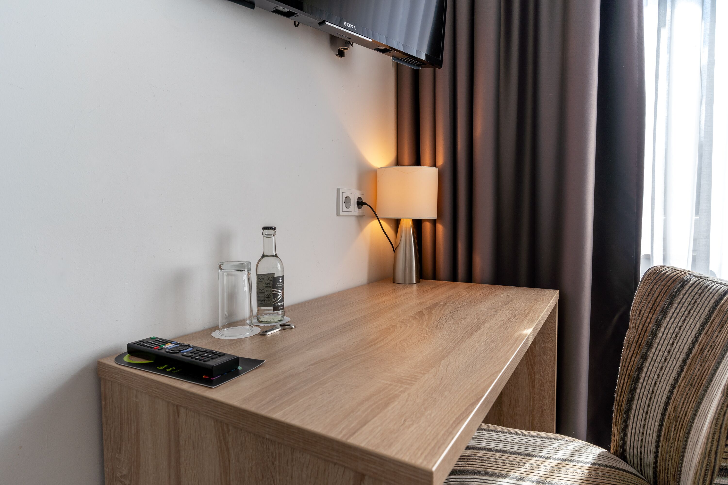 Une chambre d’hôtel avec un bureau en bois, une télécommande, une bouteille en verre et une lampe.