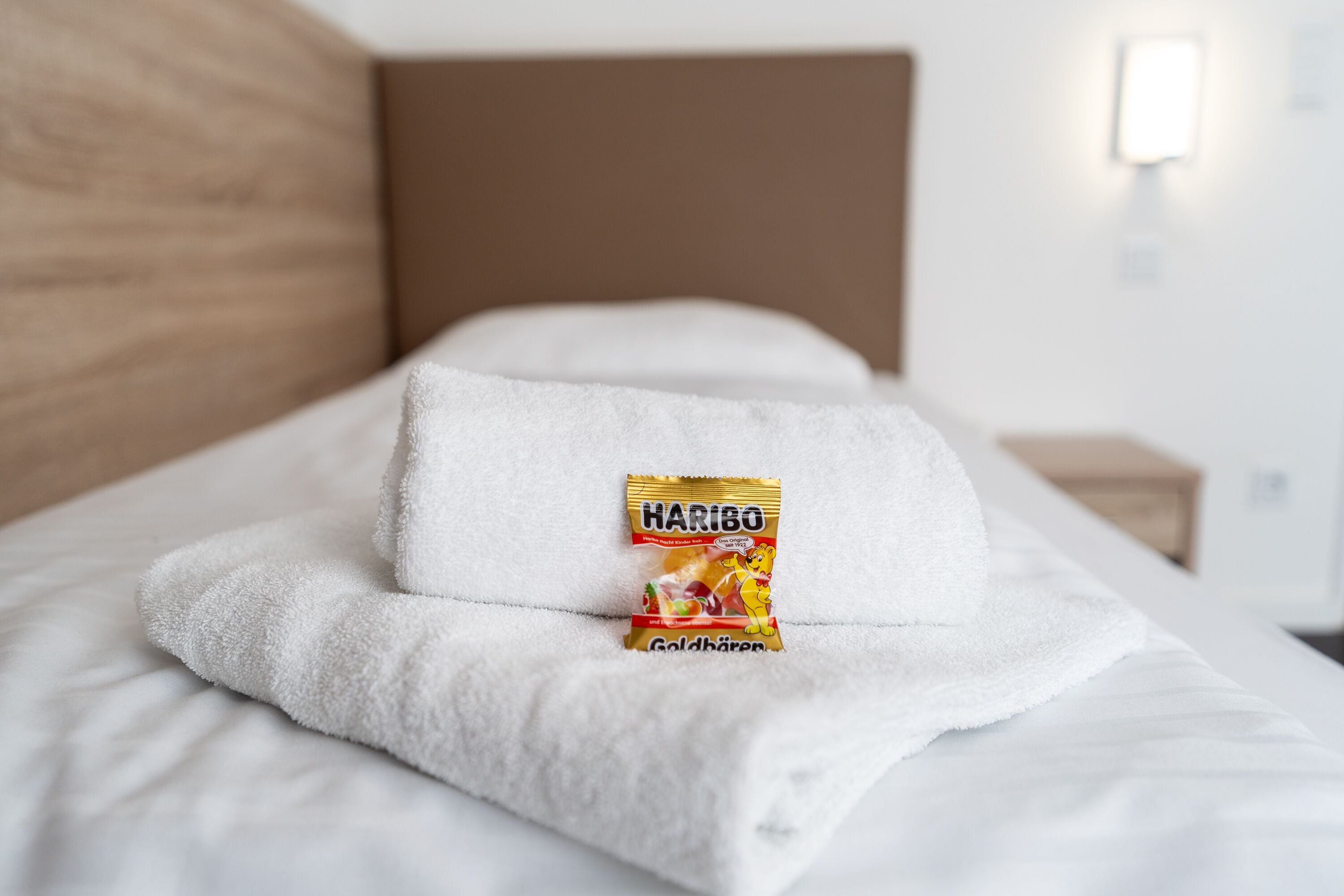 Een netjes opgemaakt bed met een witte handdoek en een zakje Haribo Goldbären erop.