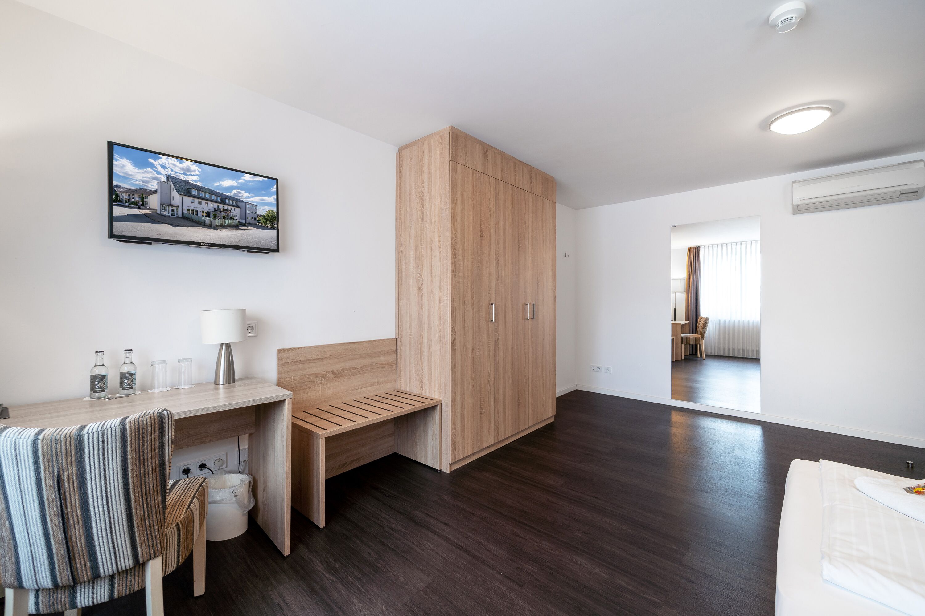 Una habitación de hotel moderna con un armario de madera, un escritorio con una silla, un banco y una cama.