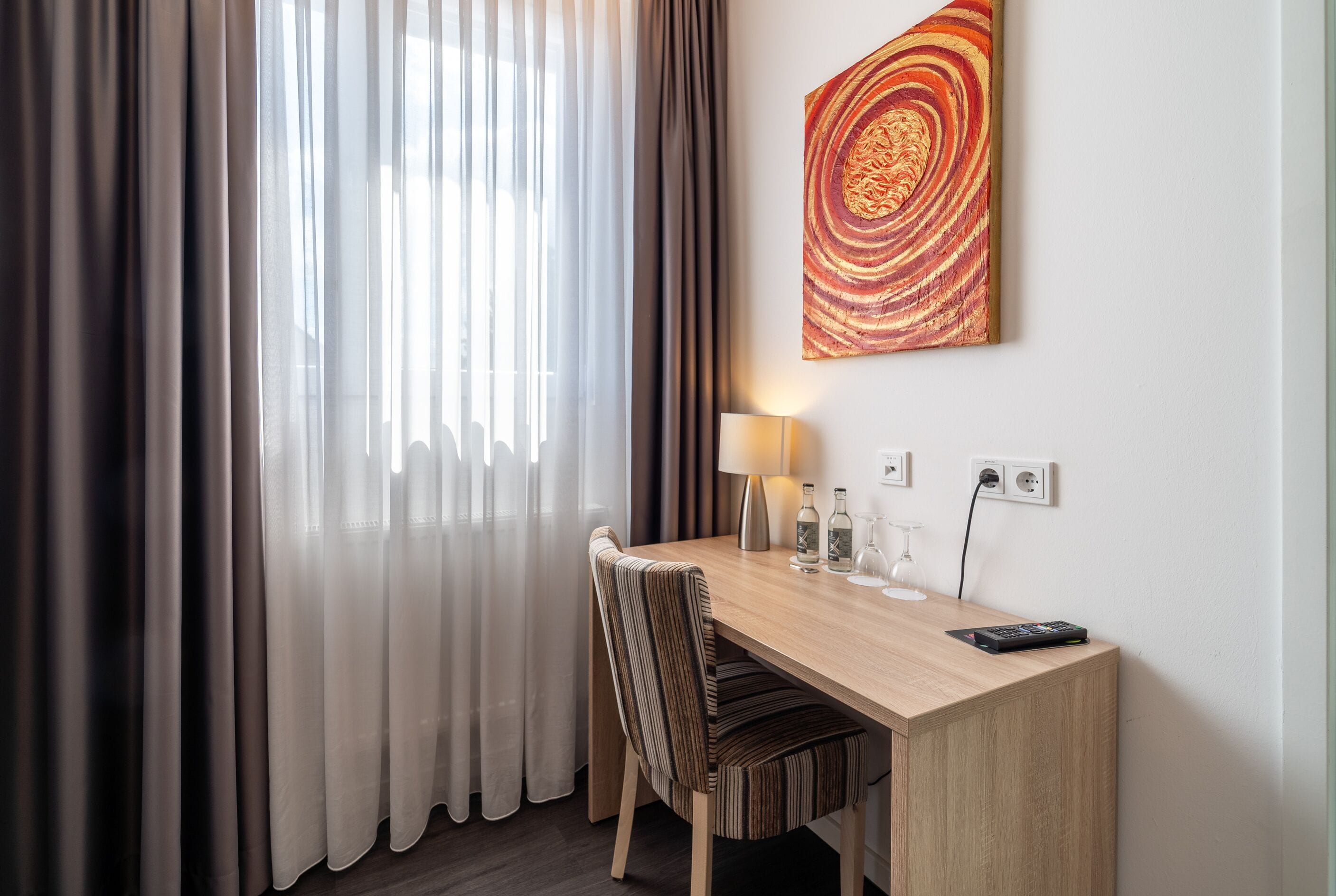 Habitación de hotel con un escritorio de madera, una silla a rayas, una lámpara, un control remoto y un cuadro en la pared.