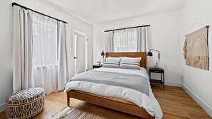 Un dormitorio con una cama de madera, dos mesitas de noche con lámparas, un pequeño reposapiés y un tapiz grande tejido.