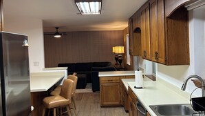 A modern kitchen with wooden cabinets, a stainless steel refrigerator, and a dining area with bar stools. - Crew-Ready Near Nine Mile Pensacola Stay (Pensacola)