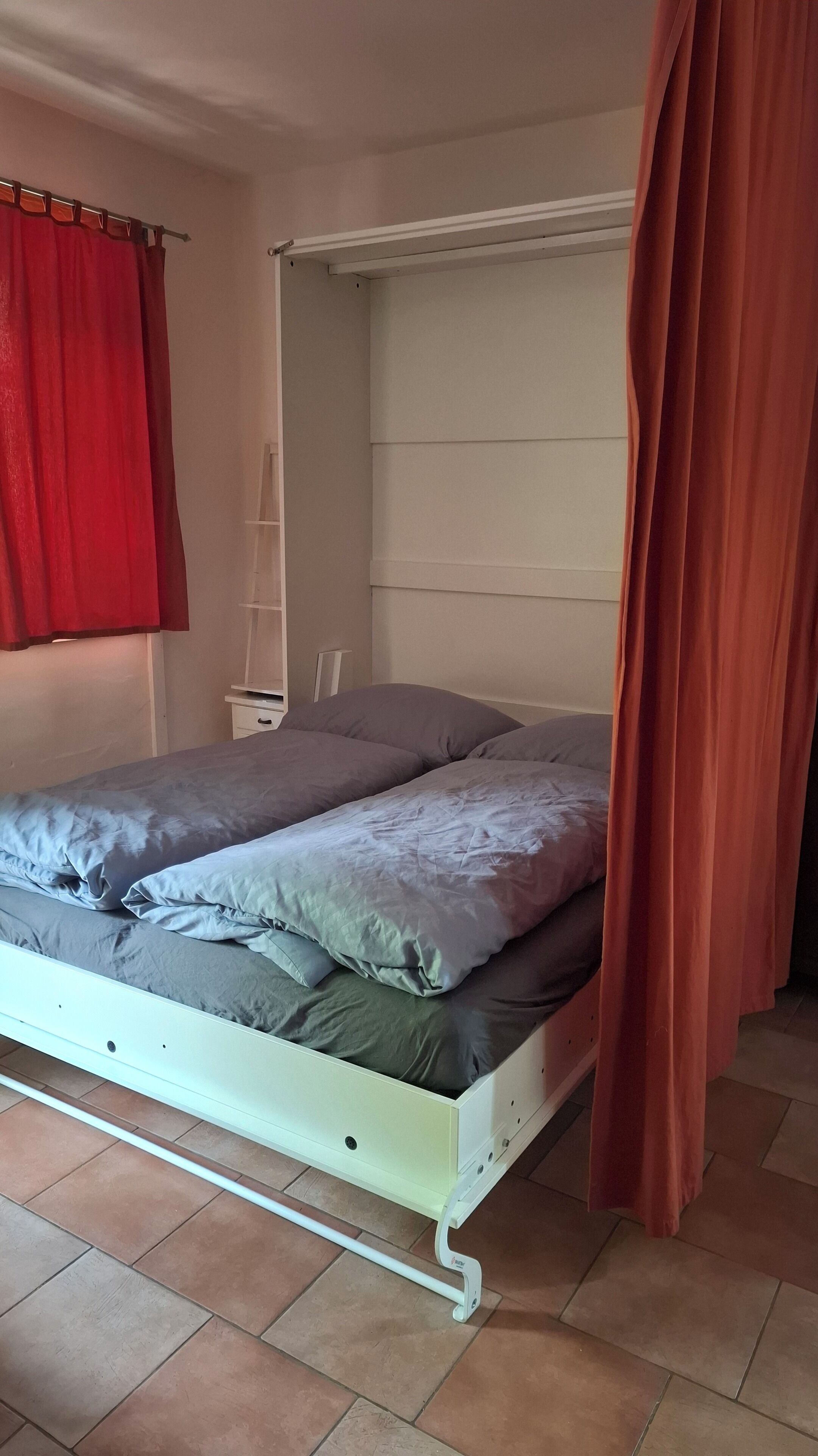 Una cama con estructura blanca, ropa de cama gris y una cortina roja entreabierta.