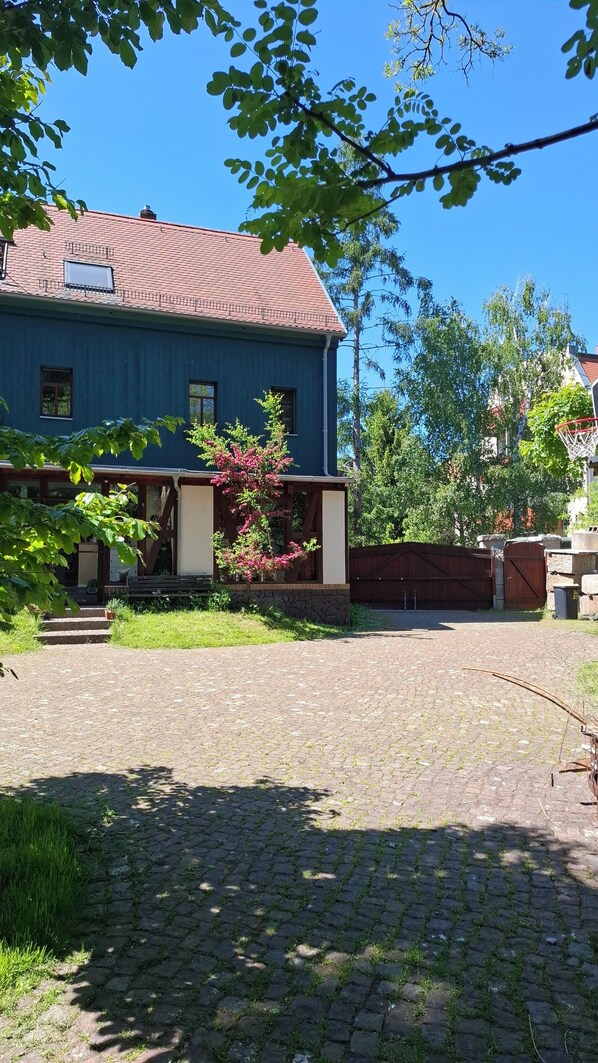 A blue house with a tiled roof, a paved driveway, and a garden area with a bench and flowering plants.