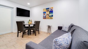 A modern living room with a grey sofa, a dining table with chairs, a small side table, and a colorful abstract painting on the wall.