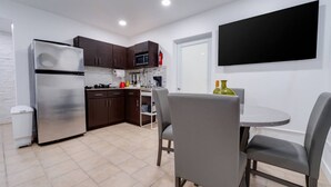 A modern kitchen with dark cabinets, a stainless steel refrigerator, and a microwave. There is a dining area with a round table and two chairs. A flat-screen TV is mounted on the wall above the table.
