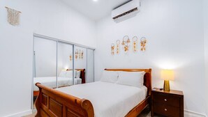 A bedroom with a wooden bed, a nightstand with a lamp, a wardrobe with mirrors, and an air conditioning unit.