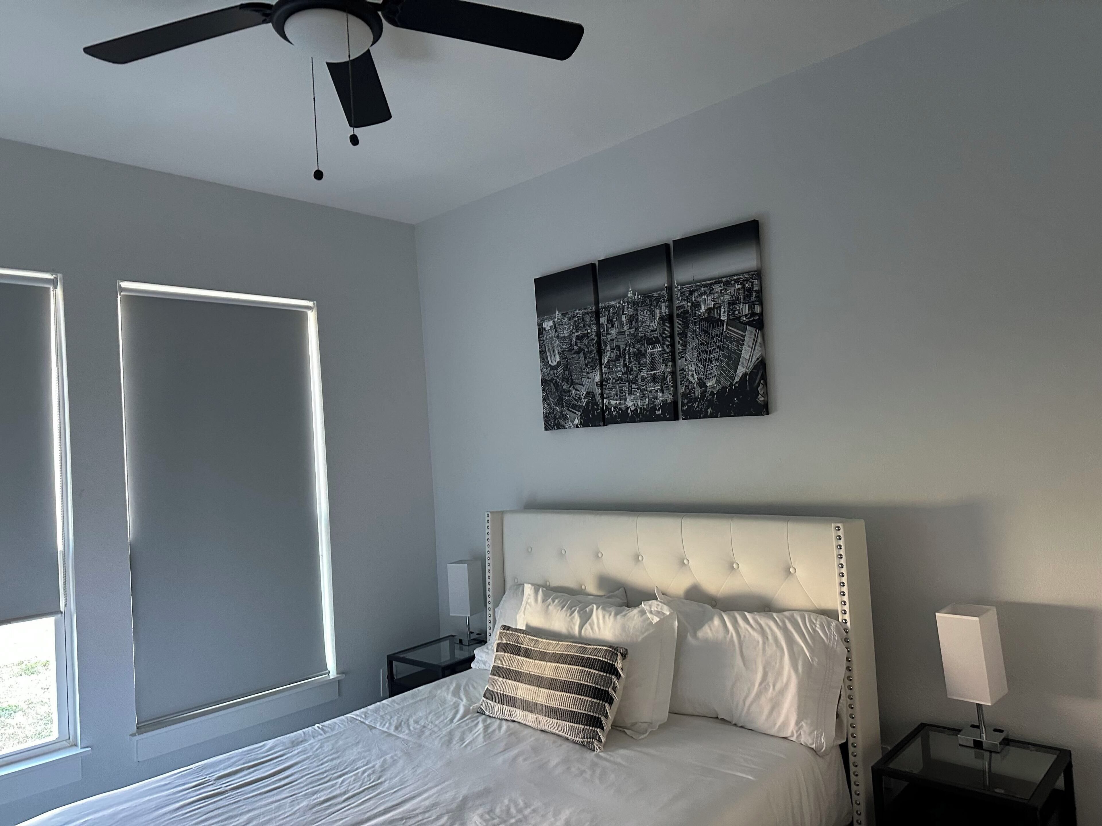 A bedroom with a bed, a ceiling fan, a nightstand, a lamp, and a cityscape artwork on the wall.