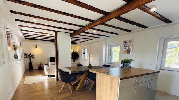 A modern kitchen with wooden beams, a central island, and a dining area with chairs. - Cozy Family Home near Ramstein Air Base – Comfort & Convenience (Hochspeyer)