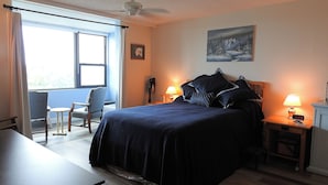 A bedroom with a bed, two bedside tables with lamps, a chair, a desk, a window with curtains, and a painting on the wall. - Seasonal Rental: 1-bedroom condo, gated community, sunrises over Old Tampa Bay (Clearwater)