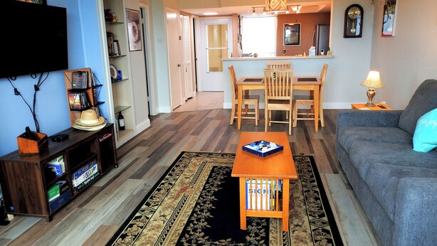 A living room with a sofa, a coffee table, a TV, and a bookshelf. There is a dining area with a table and chairs. - Seasonal Rental: 1-bedroom condo, gated community, sunrises over Old Tampa Bay (Clearwater)