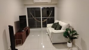 Ein Wohnzimmer mit einem weißen Sofa, einem Fernsehschrank, einem Topf mit Pflanze und einer Glastür, die auf einen Balkon führt.