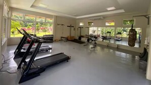 Ein großzügiger Fitnessraum mit Laufbändern, Krafttrainingsgeräten und einem Boxsack. Große Fenster sorgen für natürliches Licht und Blick auf Grünflächen.