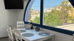 Un espace de repas moderne avec une table dressée pour deux personnes, un téléviseur fixé au mur et une vue sur les bâtiments et les arbres, visible à travers une grande fenêtre.