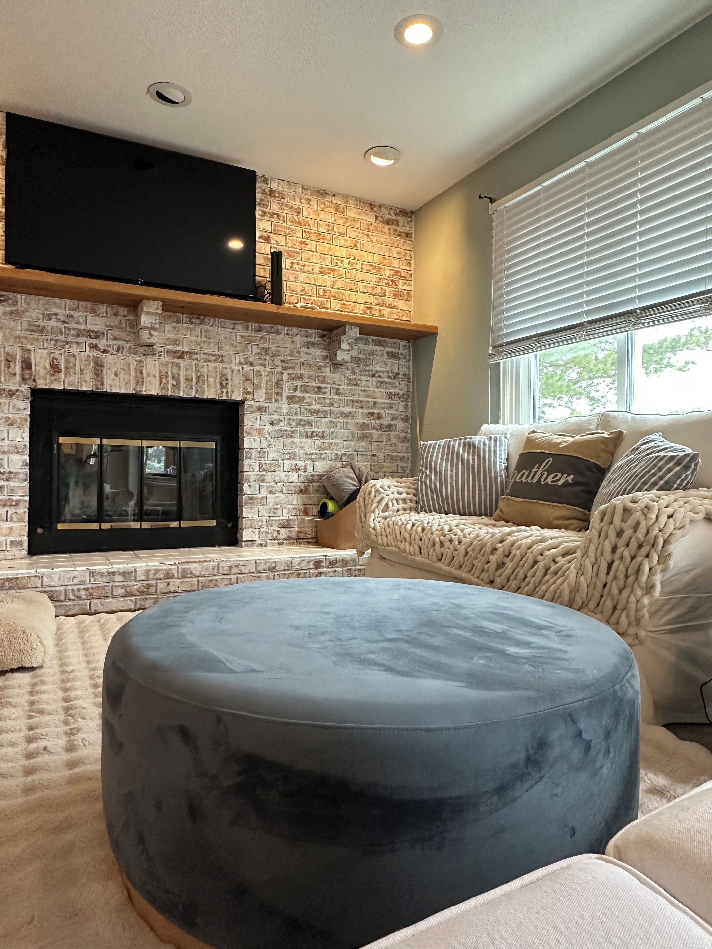 A living room with a brick fireplace, a flat-screen TV mounted above it, a beige sofa with striped pillows, and a large round ottoman.