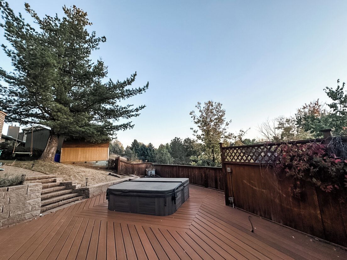 A deck with a hot tub, a wooden fence, and a large tree.
