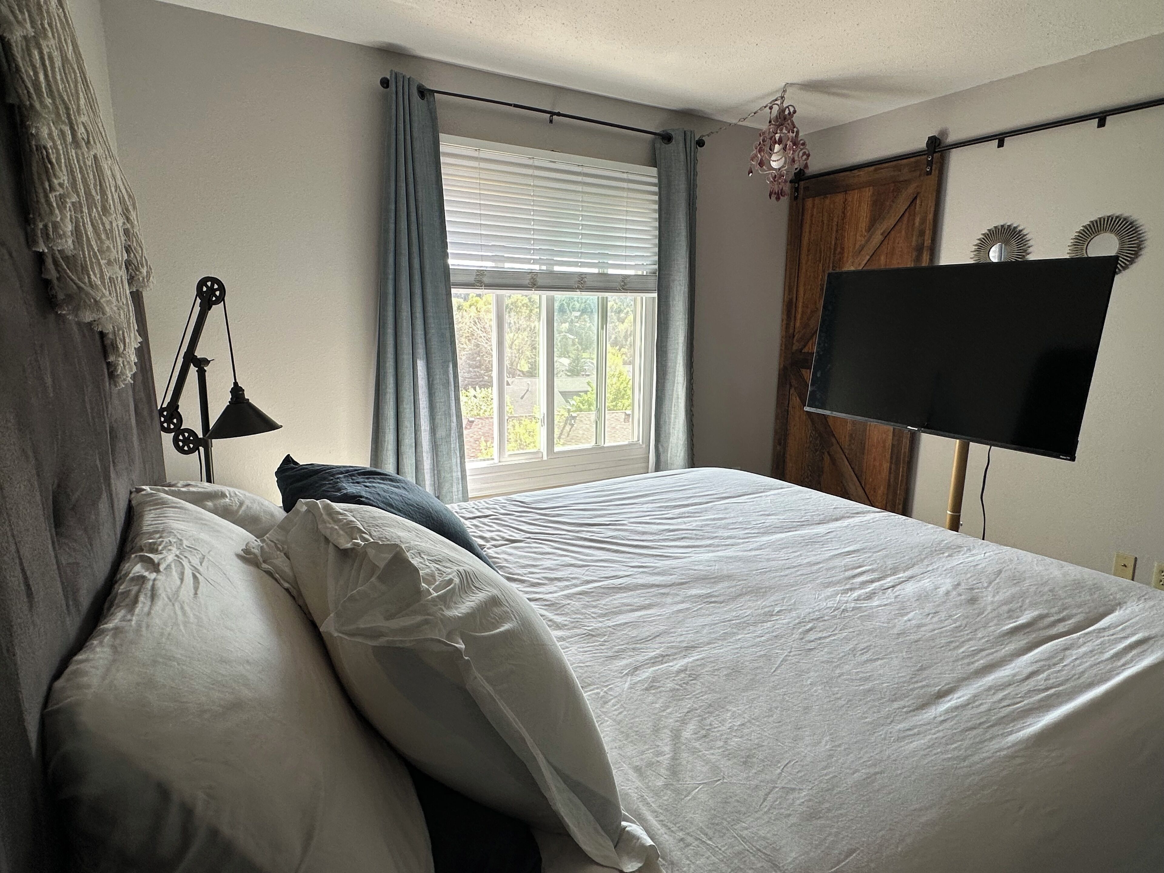 Un dormitorio con cama, ventana con cortinas, televisor montado en la pared y lámpara colgante.