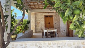 Un área de estar al aire libre con paredes de piedra, una mesa y sillas de madera, rodeada de abundante vegetación y vistas a las montañas.