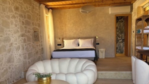Un dormitorio con pared de piedra, techo de madera, cama con ropa blanca, mesita de noche pequeña y una silla.