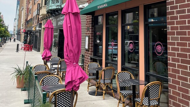 A row of outdoor cafe tables and chairs with pink umbrellas, a brick building with a green awning, and a sign reading "Rosa Blanca".